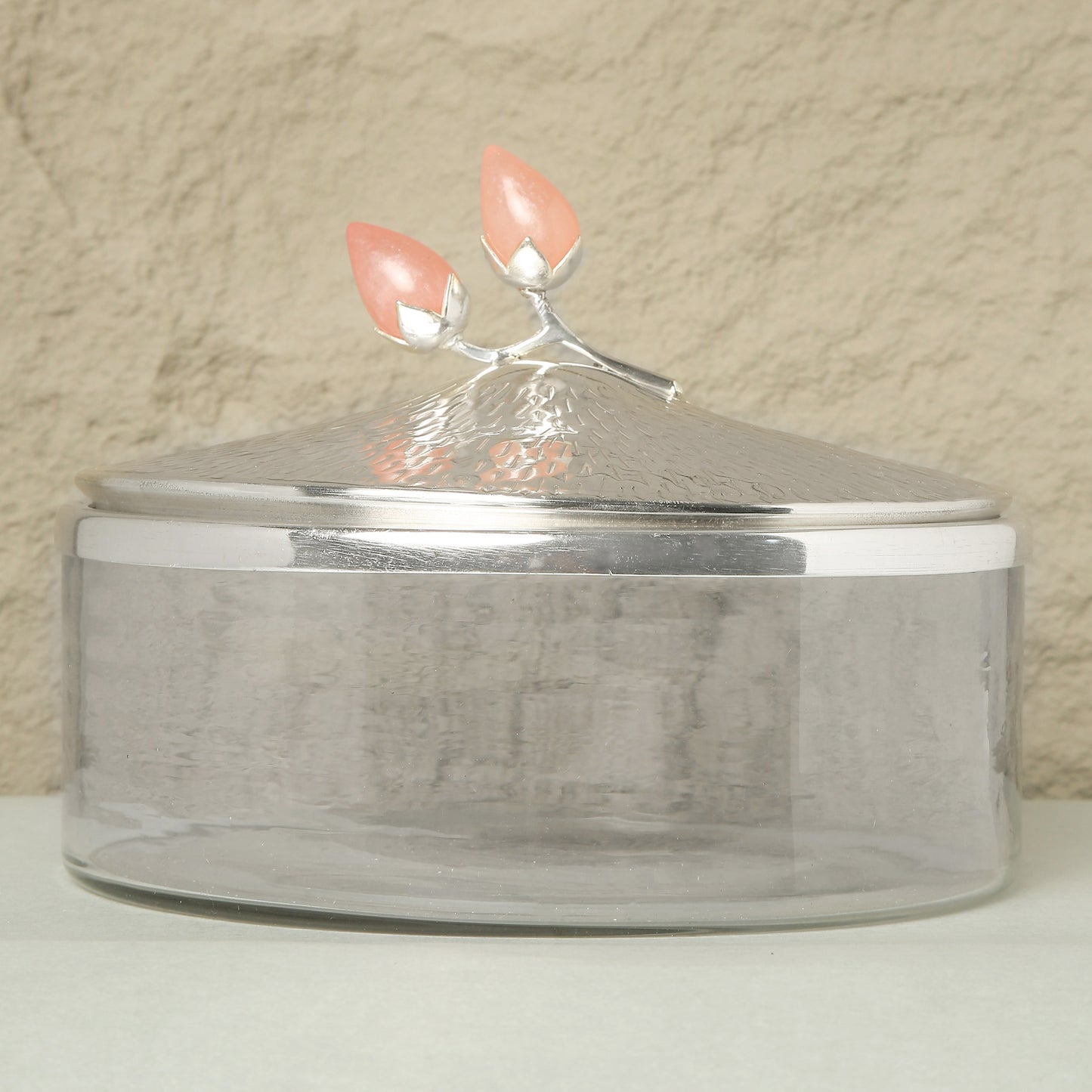 Clear glass container with a silver lid featuring coral-colored handles on a beige background