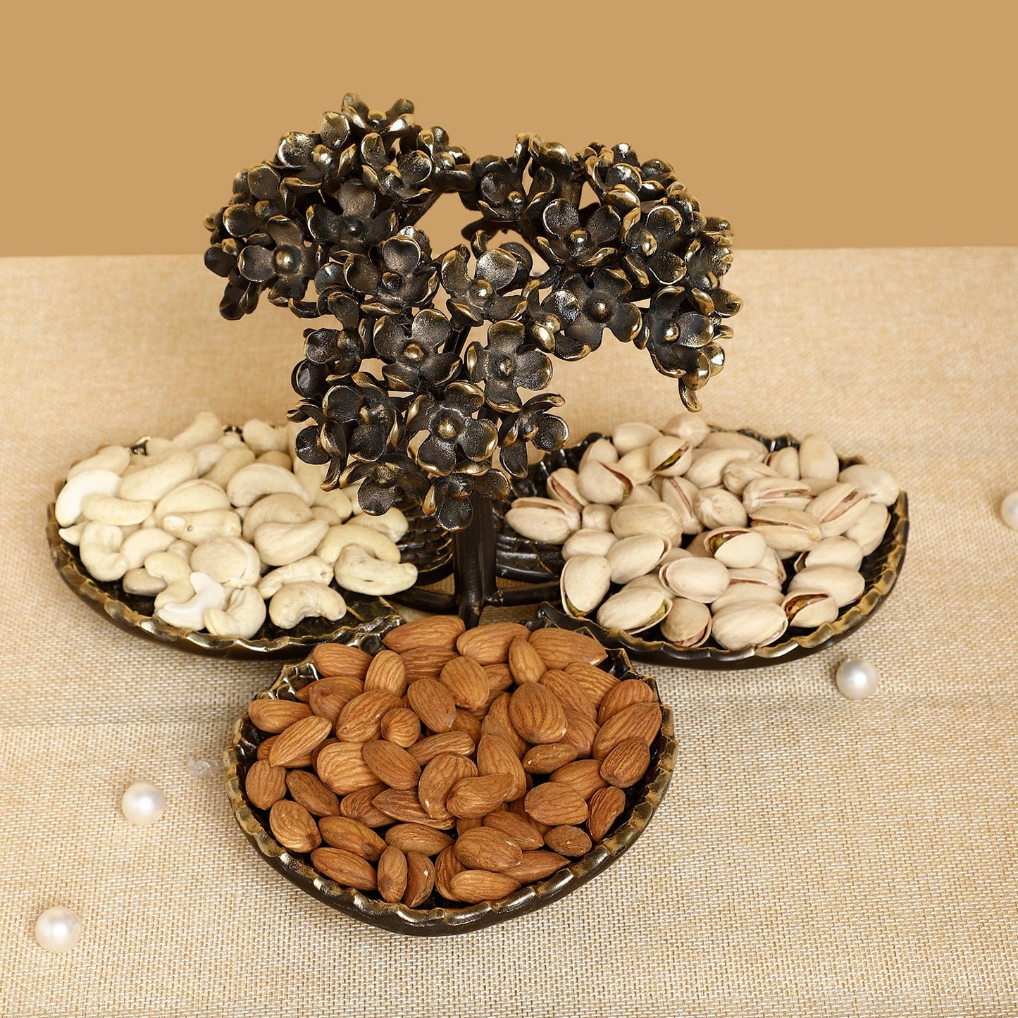 Decorative metal tree with three round trays holding almonds, pistachios, and coffee beans on a beige background.