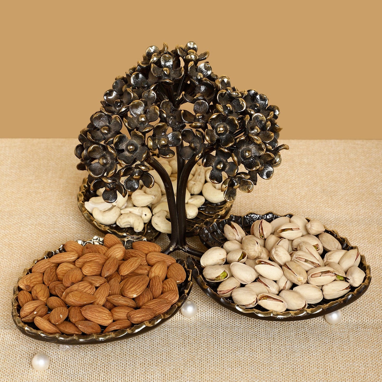 Decorative metal tree with compartments filled with almonds, cashews, and pistachios on a beige background.