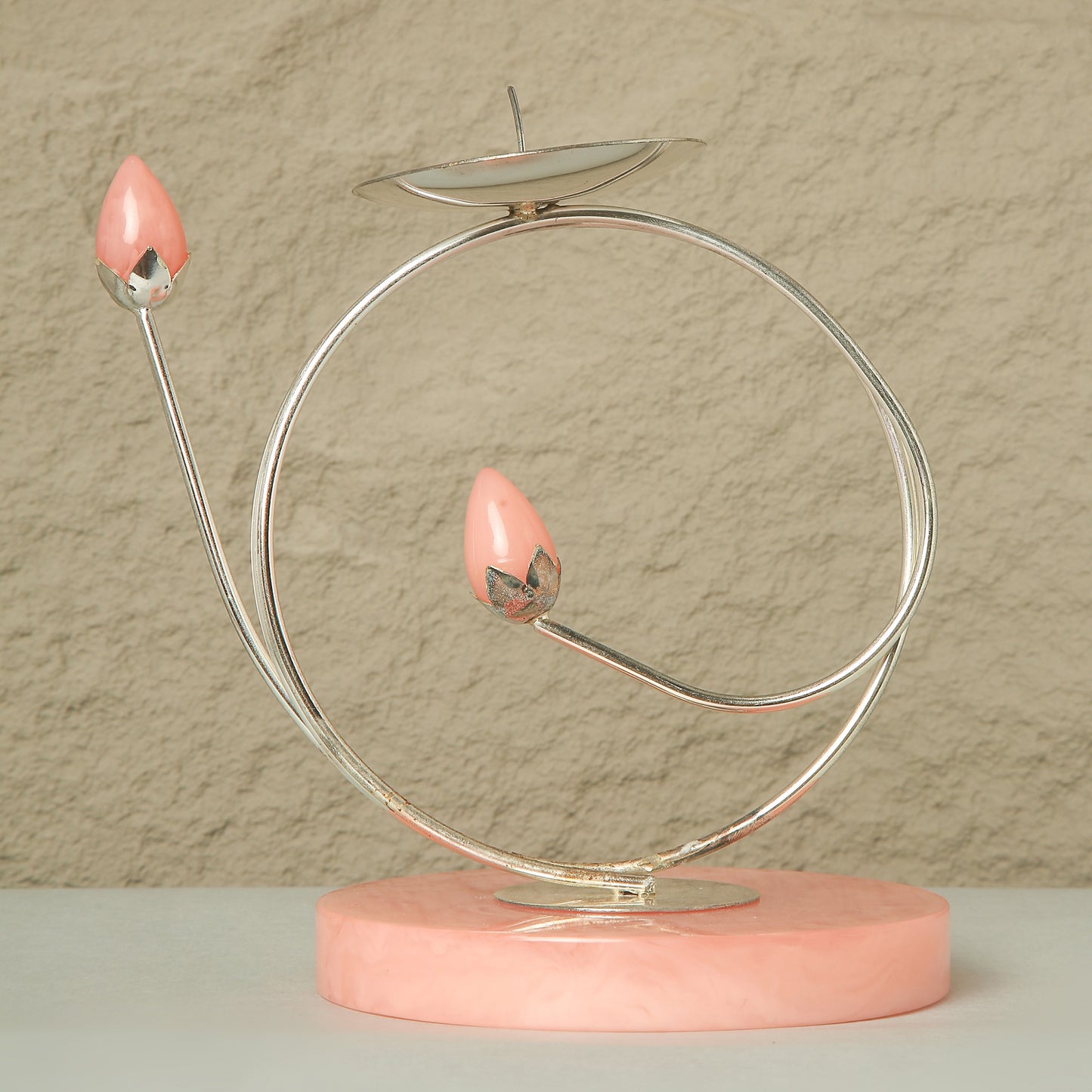 Decorative object with pink candle-like elements on a circular metal stand against a beige wall.