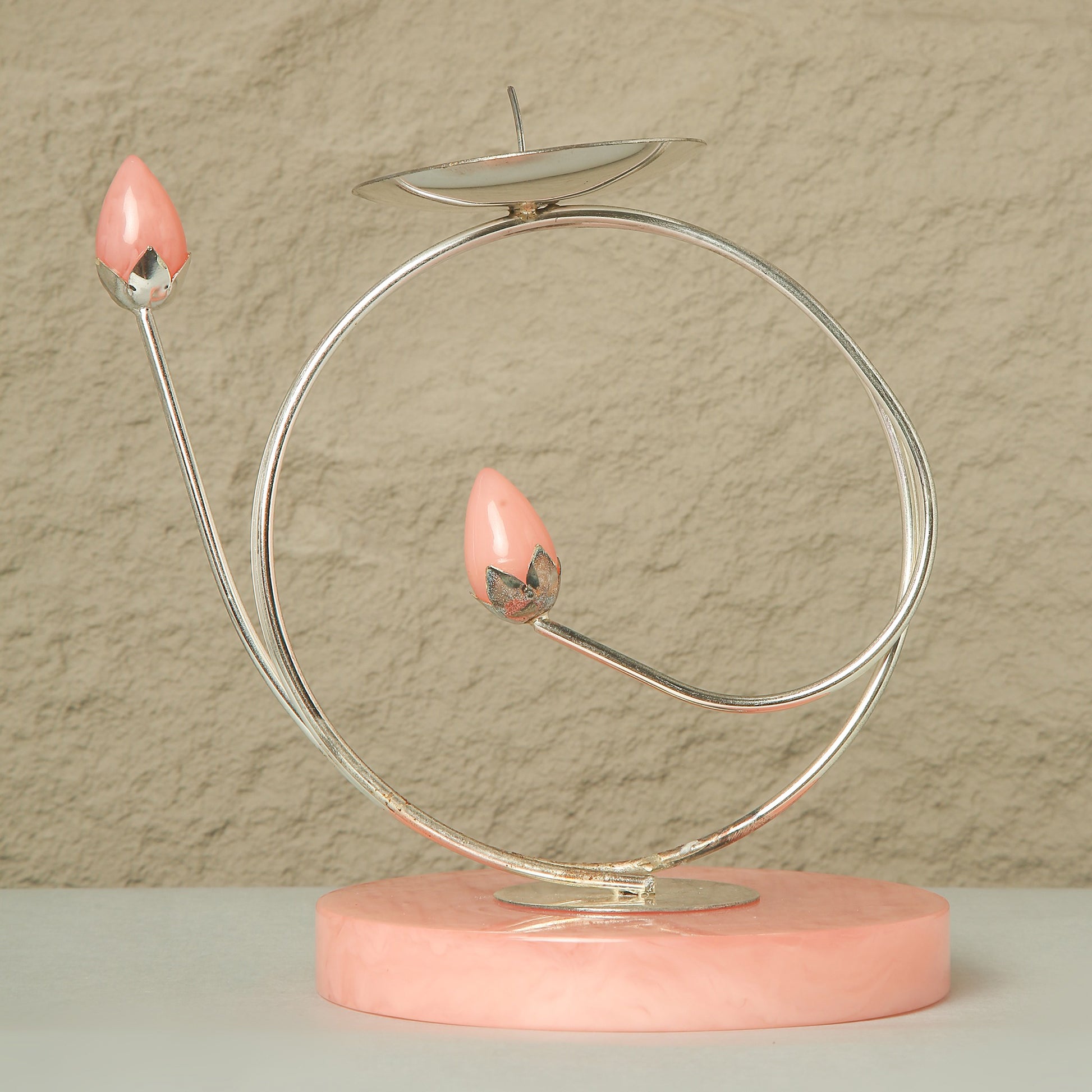 Decorative object with pink candle-like elements on a circular metal stand against a beige wall.
