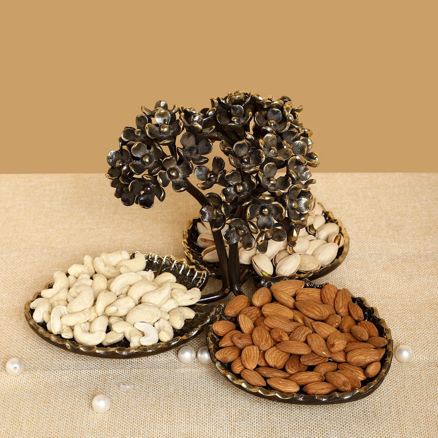 Decorative metal tree with three round trays holding almonds, cashews, and peanuts on a beige background.